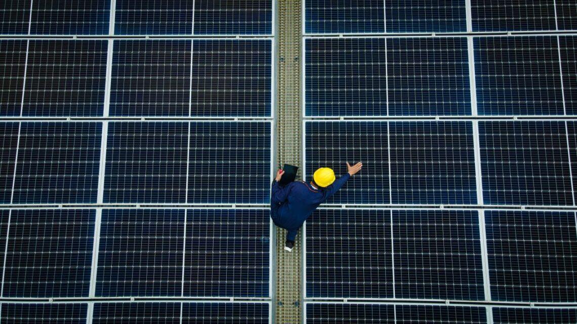 Techniker mit Helm inspiziert Glas glas photovoltaik module auf grosser Solaranlage von oben